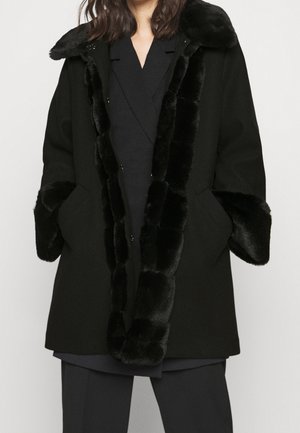 Kvinde iført en sort frakke med blød pelskant på krave, front og manchetter, hænderne i lommerne, over et sort outfit mod en ensfarvet baggrund.