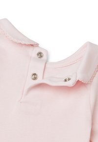 Světle růžová bavlněná košile s skládacím límcem, která má na krku dvě stříbrné patentky a detail s vlnitým okrajem na límci.