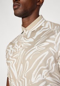 Beige Poloshirt mit abstrakten weißen Mustern, kurzen Ärmeln und einem Kragen mit gestreiftem Design. Knöpfe sind in der Nähe des Ausschnitts sichtbar.