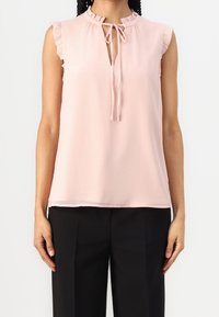 Blouse rose clair sans manches avec accents à volants, col à nouer en forme de trou de serrure, tissu doux ; associée à un pantalon noir. Design élégant et simple.