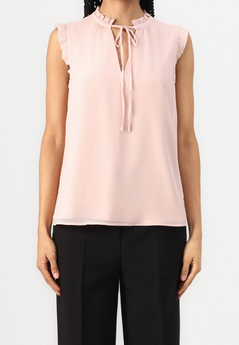 Blouse rose clair sans manches avec accents à volants, col à nouer en forme de trou de serrure, tissu doux ; associée à un pantalon noir. Design élégant et simple.