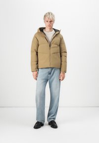 Puffer jacket in khaki with a hood, zip closure, and quilted design. Paired with light blue loose-fitting jeans and black shoes.