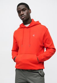 Piros pamut kapucnis pulóver húzózsinóros kapucnival, elülső zsebbel és hímzett logóval a bal mellrészen. Szürke nadrággal viselve, amely oldalzsebbel rendelkezik.