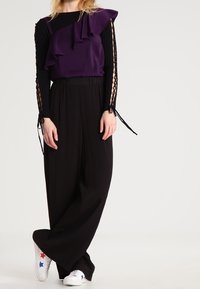 Top viola con rouches, pantaloni neri a gamba larga e sneakers bianche con stelle rosse e blu. Le maniche lunghe presentano dettagli con lacci.