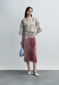 Femme portant un haut transparent brodé de fleurs, une jupe midi plissée rose avec des détails floraux, des talons blancs, et tenant un sac à main bleu clair.