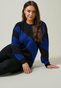 Pull rayé noir et bleu avec des manches longues, un col rond et une texture douce, porté sur un pantalon noir. Présente des motifs diagonaux audacieux.