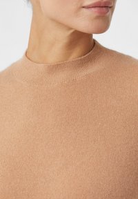 Nahaufnahme einer Person, die einen beigen Rippstrickpullover mit Stehkragen trägt, wobei der Hals und der untere Teil des Gesichts zu sehen sind.