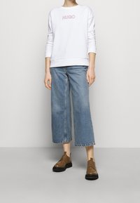 Witte sweatshirt met het 'HUGO' logo in roze, gecombineerd met lichtblauwe jeans met wijde pijpen en bruine veterschoenen met zwarte zolen.