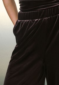 Juodos aksominės kelnės su surinktąja juosmens juosta ir šoninėmis kišenėmis, su lygiu paviršiumi ir laisvu siluetu, pritvirtintos ties klubais.