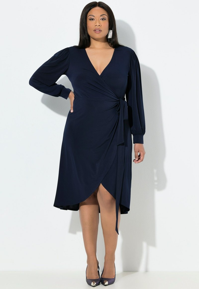 Marineblauwe wikkeljurk met lange mouwen, diepe V-hals, geplooide schouders, zijstrikdetail en een asymmetrische zoom tot de knie. Zachte, gladde stof.