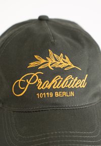 Dark green baseball cap with yellow embroidered text "Prohibited 10119 Berlin" and a leaf design above the text.
