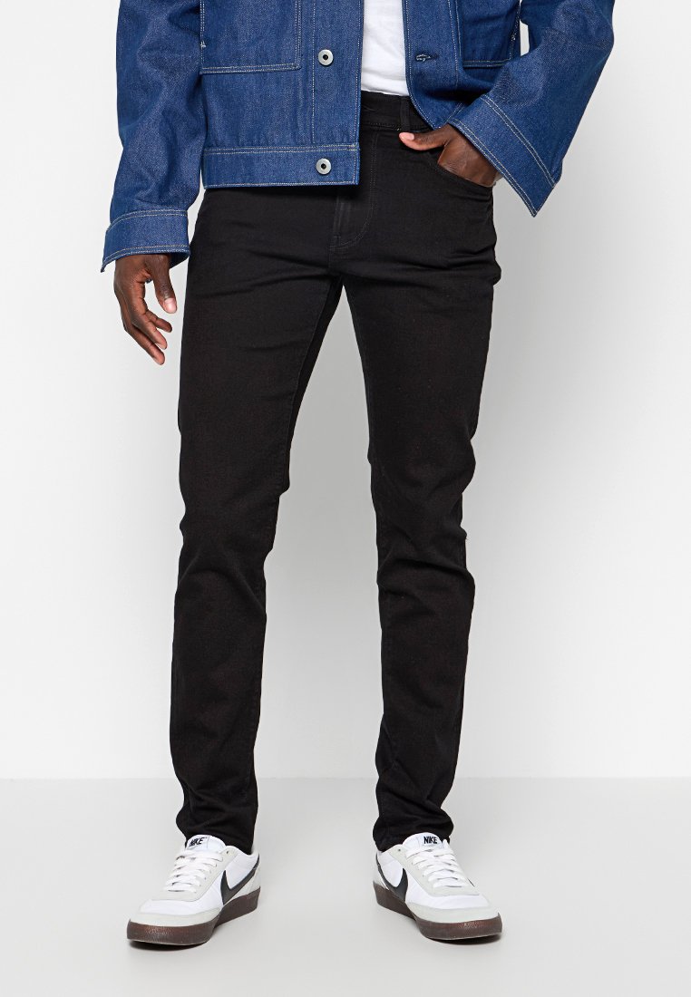 Jean en denim noir avec une coupe slim, associé à une veste en denim bleu et des baskets blanches. Présente un couture classique et un design de poches avant.