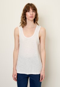 Balta, be rankovių tankų marškinėliai su iškirpte, pagaminti iš lengvos medžiagos, turinčios šiek tiek permatomą tekstūrą, derinami su mėlynais džinsais.