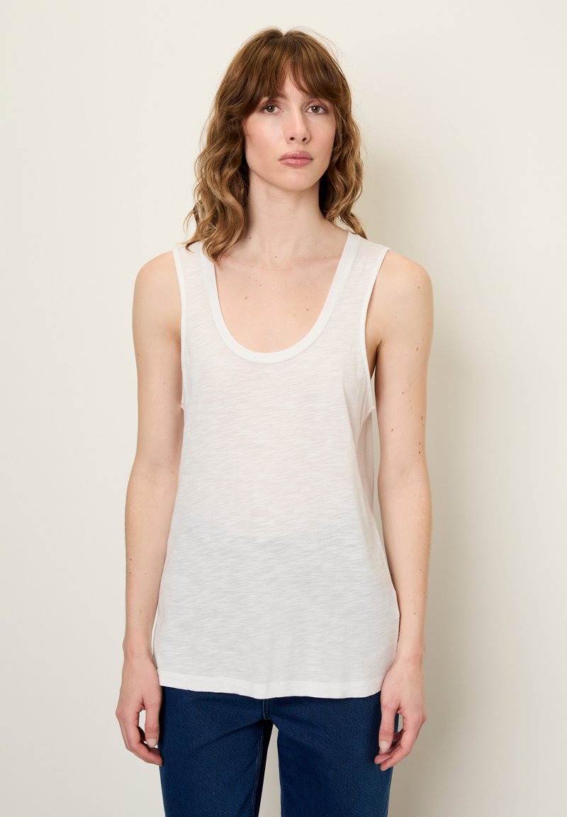 Balta, be rankovių tankų marškinėliai su iškirpte, pagaminti iš lengvos medžiagos, turinčios šiek tiek permatomą tekstūrą, derinami su mėlynais džinsais.