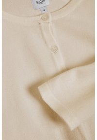 Beige kofta i mjukt tyg, med rund halsringning, knappstängning och ribbade muddar, med en etikett som visar "Saint Tropez" (M).