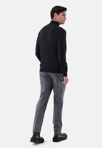 Schwarzer gestrickter Pullover mit hohem Kragen, kombiniert mit grauen gemusterten Hosen. Das Modell steht mit dem Rücken zur Kamera und trägt schwarze Stiefeletten.