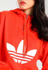 Red athletic jacket with a hood, featuring a large white Adidas logo and stripe design on the front. Smooth texture and lightweight material.