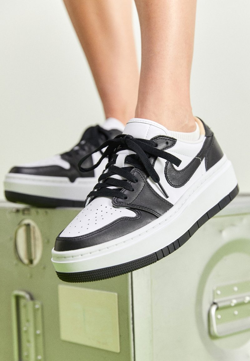 Black and white low-top sneakers feature leather upper, perforated toe, black laces, and rubber sole with textured detailing.