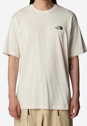 Persoon draagt een losse, off-white The North Face T-shirt met korte mouwen en beige broek, toont een kleine zon-tatoeage op de linker onderarm.