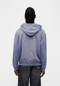 Μωβ φούτερ μ ε κουκούλα και κορδόνι, άνετη γραμμή και ριμπ τελείωμα στα μανίκια, συνδυασμένο με σκούρο τζιν, εμφανίζοντας λεπτομέρειες στην πλάτη.