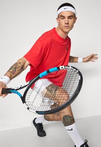 Punainen puuvillapaita, valkoiset shortsit, mustat tennarit, valkoinen hiuspanta. Pitää mustaa ja sinistä tennismailaa, esittelee yksityiskohtaisia tatuointeja käsivarsilla.