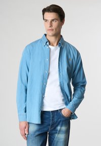 Camicia button-up azzurro chiaro in denim, con un design casuale, colletto a punta e un piccolo logo sul petto, abbinata a jeans blu.