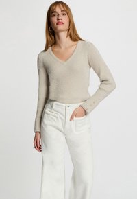 Mujer vistiendo un suéter de pico beige de peluche con botones dorados en las mangas y unos pantalones blancos de pierna ancha y cintura alta, posando con una mano en el bolsillo.