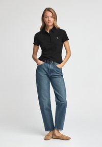 Camisa polo negra de manga corta con cuello y tres botones, combinada con jeans rectos de mezclilla azul claro y zapatos slip-on beige.