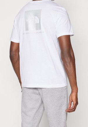 Mies, jolla on valkoinen The North Face -t-paita, jossa on logo ja teksti "Never Stop Exploring" takana, yhdistettynä harmaisiin verkkareihin.