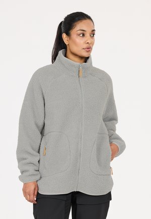 Frau mit dunklen Haaren im Pferdeschwanz, die eine hellgraue Fleecejacke mit Reißverschluss und Taschen trägt und vor einem einfachen weißen Hintergrund steht.