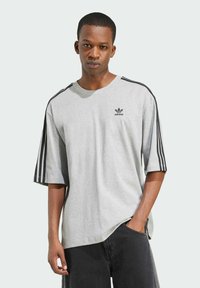 Maglietta oversize grigia chiara con accentuazioni nere a tre strisce sulle spalle e piccolo logo nero sul petto. Materiale in cotone, maniche corte.