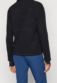 Giacca in pile nera con colletto alto, caratterizzata da cuciture piatte e maniche lunghe, abbinata a pantaloni blu per un contrasto.