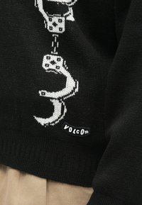 Vêtement tricoté noir avec un graphique blanc de menottes et l'étiquette de la marque "Volcom" près de l'ourlet, au-dessus d'un pantalon beige.