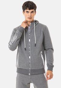 Grauer Kapuzenpullover mit weißen Kordeln, Reißverschlusstaschen, logobezogenem Detail an den Ärmeln und kontrastierendem schwarzem Band auf der Vorderseite.