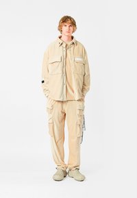 Camisa de pana beige con dos bolsillos en el pecho, combinada con pantalones cargo a juego que cuentan con múltiples bolsillos y una etiqueta. Usada con zapatos texturizados.