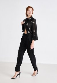 Chemise noire transparente à boutons avec des motifs floraux brodés, associée à un pantalon noir ample et des sandales à talons noires avec des embellissements.
