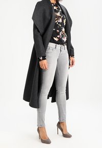 Manteau en laine gris avec un col large, blouse noire à motifs floraux, jean skinny gris clair et chaussures à talons hauts grises. Tissus texturés et design en couches.