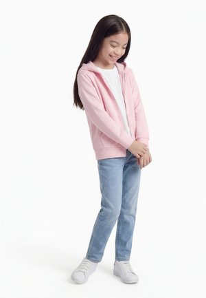 Jeune fille debout souriante, portant un sweat à capuche rose à fermeture éclair, un t-shirt blanc, un jean bleu clair et des baskets blanches, sur un fond blanc uni.