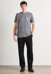Grijze katoenen t-shirt met een kleine graphic op de linkerborst, gecombineerd met zwarte losse broek en zwarte sneakers.