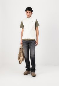 Weißer Fleece-Weste über einem grünen T-Shirt, dunklen Jeans und braunen Schuhen. Trägt eine hellbraune Tasche. Neutraler Hintergrund.