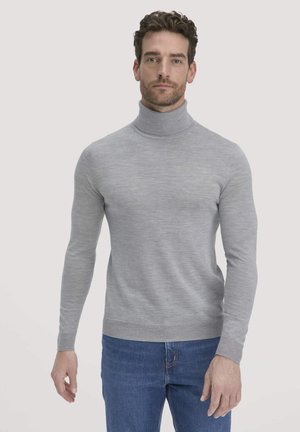 Man in een lichtgrijze coltrui en blauwe jeans, staand tegen een effen lichte achtergrond.