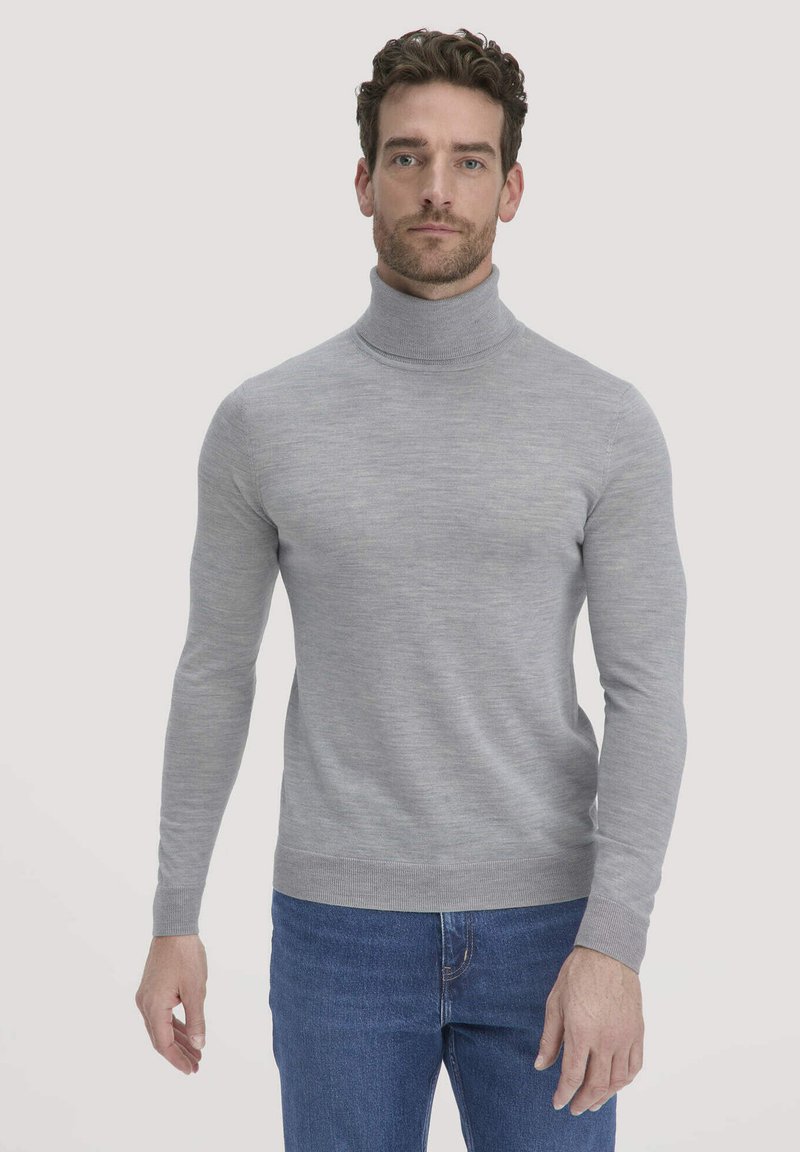 Mann in einem hellgrauen Rollkragenpullover und blauen Jeans, der vor einem einfarbigen hellen Hintergrund steht.
