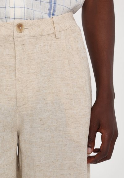 Pantalons en lin beige avec une finition texturée, dotés d'une fermeture à bouton et de poches latérales. Main visible tenant la taille.