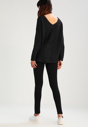 Pullover nero a maglia con design a V sul retro, abbinato a leggings neri e sneakers bianche. La texture sembra morbida e dal fit comodo.