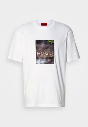 Hvit bomulls-T-skjorte med et rektangulært grafisk trykk med teksten "HUGO" på en teksturert, mørk overflate, omgitt av grøntområder.