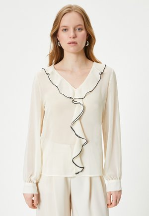 Blusa crema con escote en V, que presenta un frente con volantes y ribete negro. Mangas largas con puños abotonados, hecha de tela ligera y transparente.