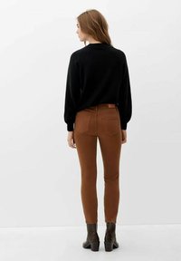Frau mit langen hellbraunen Haaren, die einen schwarzen Pullover, braune enge Hosen und dunkle Stiefeletten trägt, steht mit dem Rücken zu einem schlichten weißen Hintergrund.