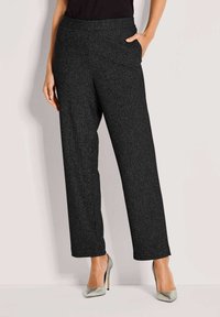 Zwarte gestructureerde wijde broek gedragen met zilveren hoge hakken, één hand in de zak, onderste helft van een persoon zichtbaar tegen een effen achtergrond.