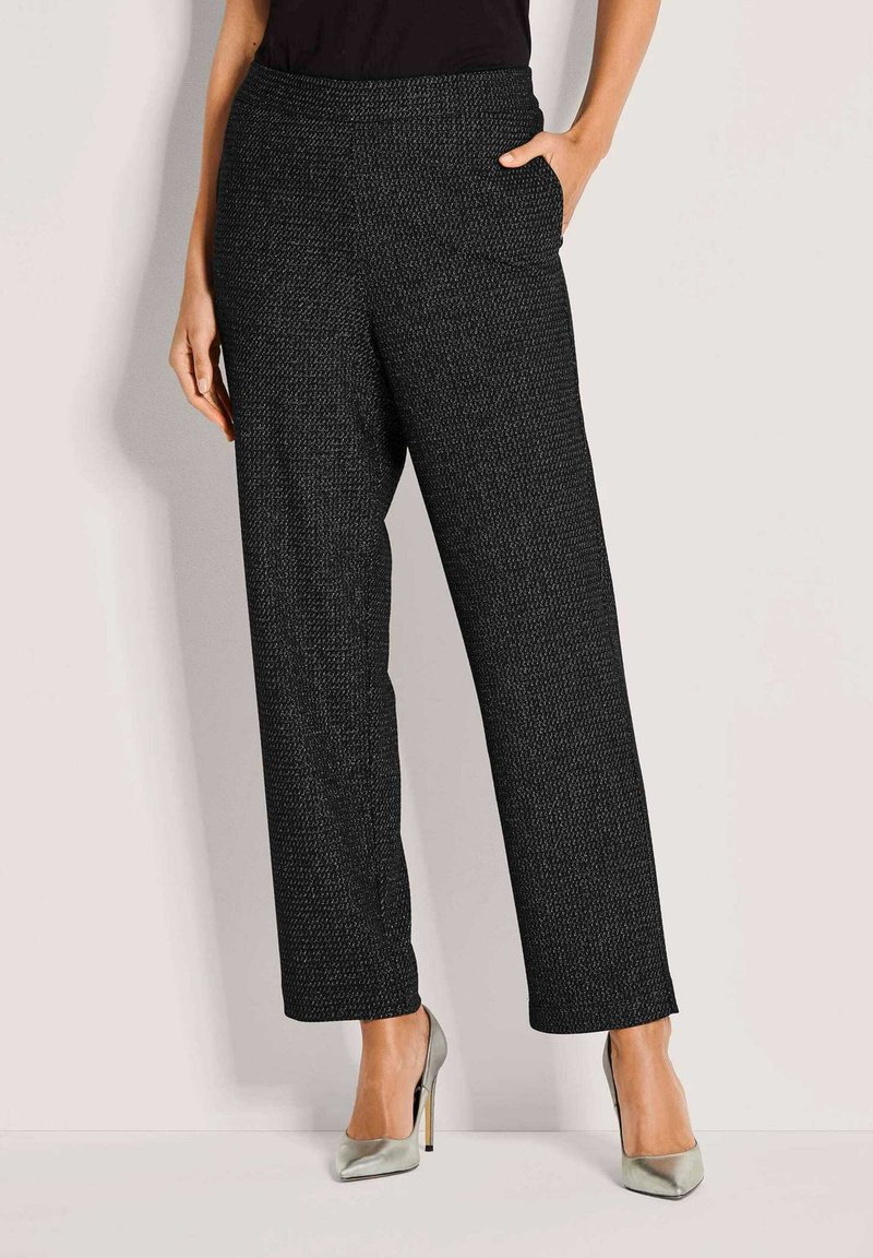 Zwarte gestructureerde wijde broek gedragen met zilveren hoge hakken, één hand in de zak, onderste helft van een persoon zichtbaar tegen een effen achtergrond.