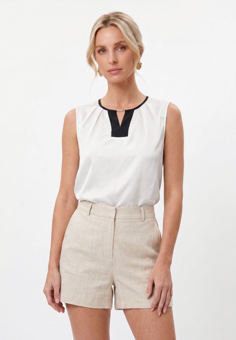 Femme blonde portant un chemisier blanc sans manches avec un détail noir à l'encolure et un short beige taille haute, debout sur fond blanc.